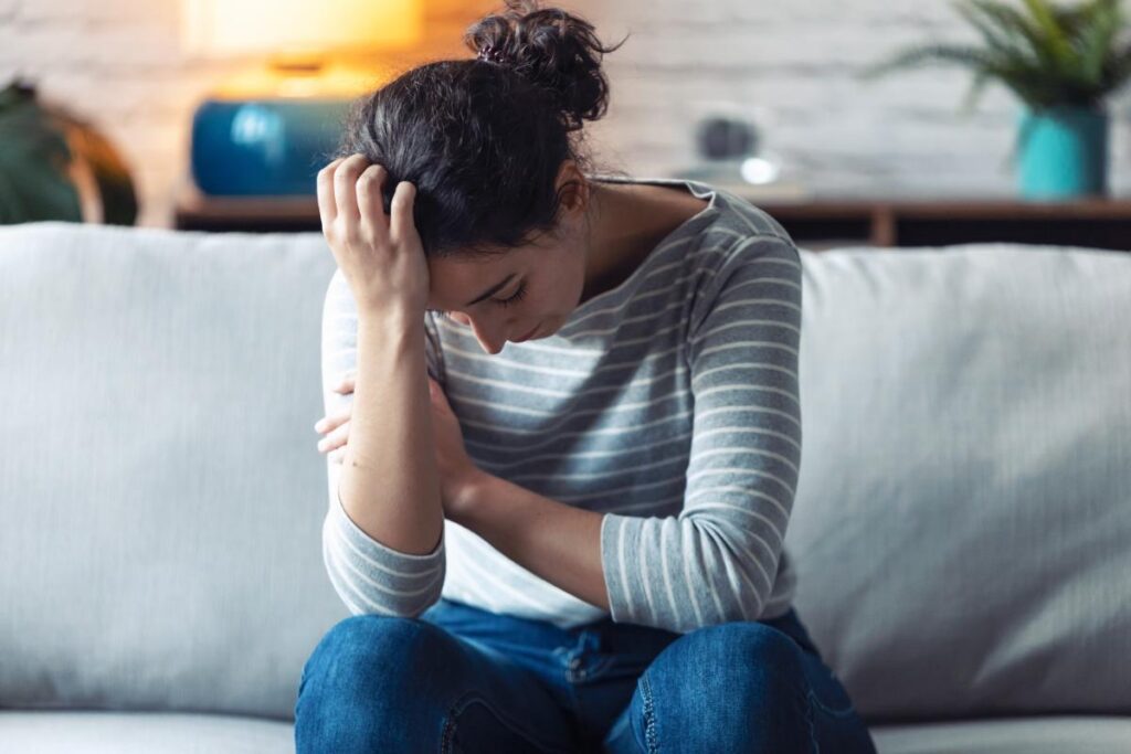 A woman has experienced an emotional trauma trigger. She sits on a couch with her head in her hands.