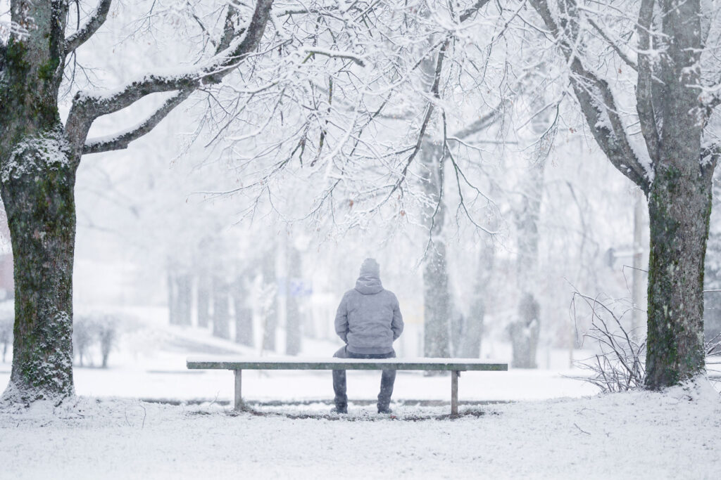 A man sits on a bench outside in the snow. He is alone and experiencing seasonal depression.