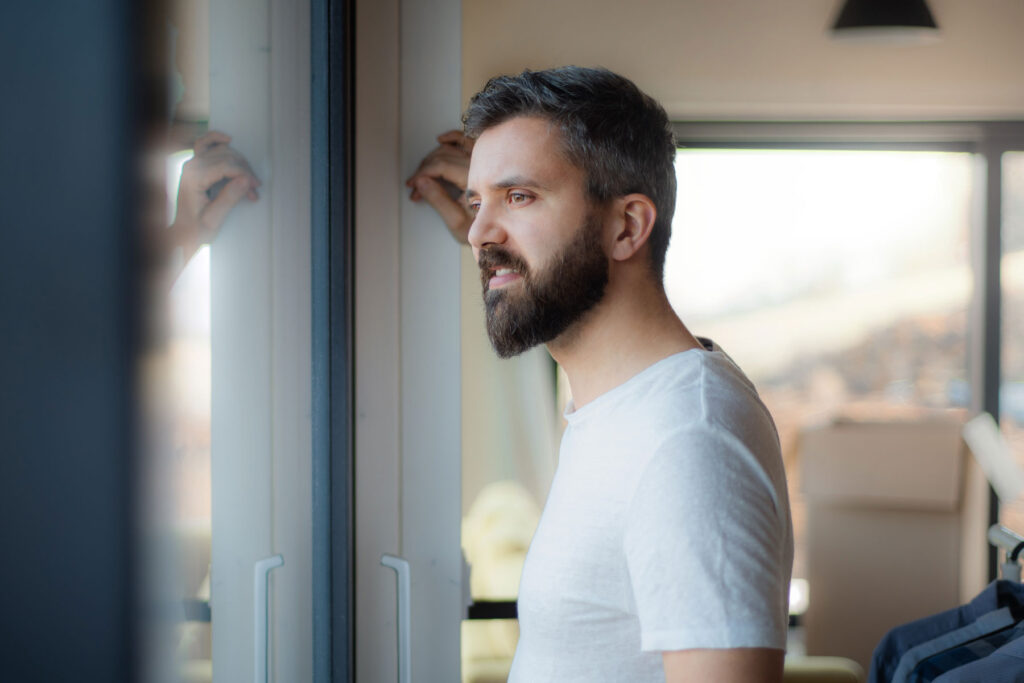 Young man standing in his home in a white tee shirt staring uneasily into space and wondering is CBD addictive?