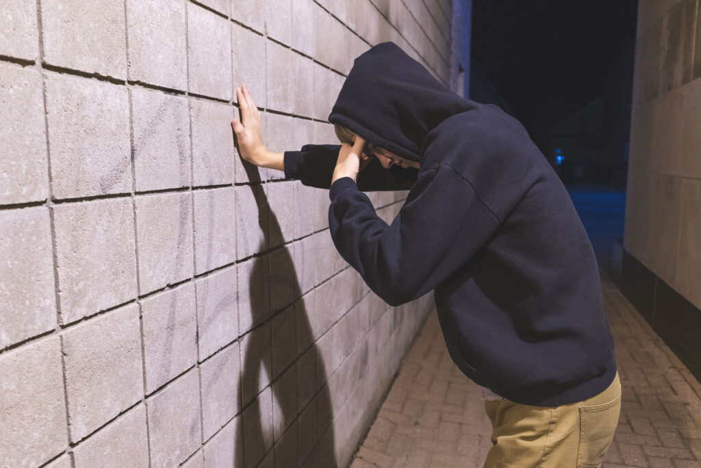 A man with a hood pulled up over his head is standing in an alley. His hand is pressed against a brick wall, while the other is at his forehead. He is experiencing the effects of alcohol and cocaine.