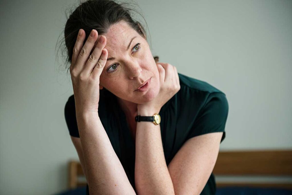 A woman in a black shirt is sitting down with one hand at her forehead and another on her neck. She is experiencing the long-term effects of alcohol.